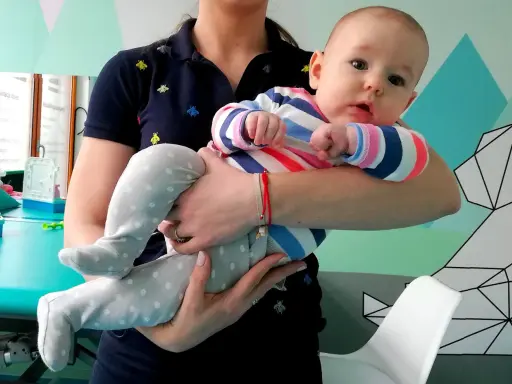 Dziecko w kolorowym ubranku, trzymane przez opiekunkę, przyjmuje pozycję amfibii. W tle widać elementy rehabilitacyjne.