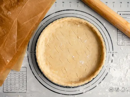 Pâte sucrée noisette prête à être garnie, sur un tapis de pâtisserie avec un rouleau à bois et du papier sulfurisé.