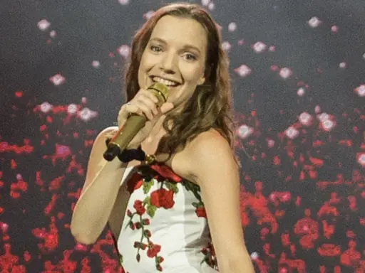 Uśmiechnięta wokalistka w białej sukience z czerwonymi różami śpiewa do złotego mikrofonu. To tata sanah na scenie.