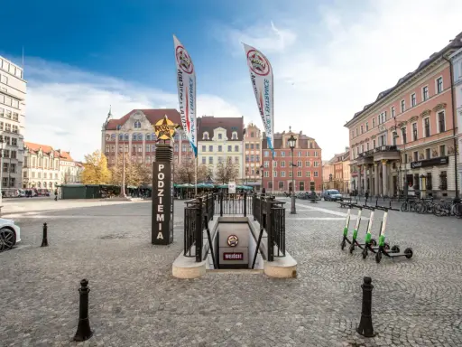 Wejście do podziemi z napisem "Movie Gate" i dwoma pionowymi banerami. W tle kolorowe kamienice i błękitne niebo.