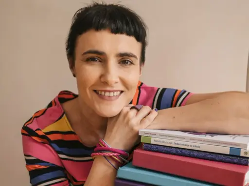 Uśmiechnięta kobieta z krótkimi włosami opiera się o stos kolorowych książek. Wiedza, którą posiadają, jest dla niej dostateczna.