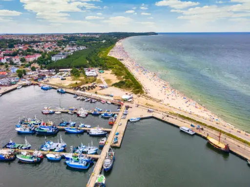 Władysławowo co zobaczyć: port z łodziami, szeroka plaża pełna turystów i miasto w tle. Idealne miejsce na wakacje nad morzem.