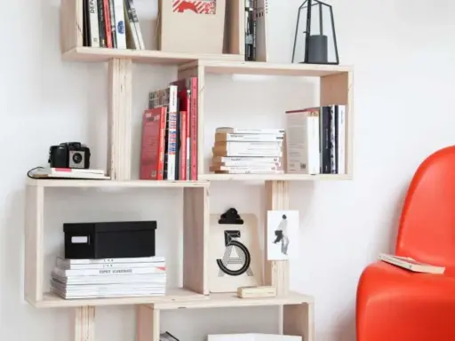 Bibliothèque DIY en bois clair, remplie de livres, d'une caméra vintage et d'une boîte noire. Une chaise rouge vif est visible à droite.