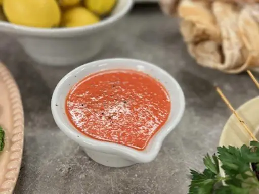 En skål med en röd, kryddig sås, kanske en pico de gallo, står på ett grått bord. Bredvid syns gula citroner och bröd.