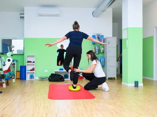 Kobieta ćwiczy propriocepcję na poduszce balansującej, druga osoba asekuruje.