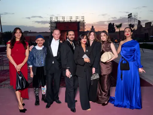 A group poses at a fashion event in Arabia. The backdrop features a large screen with "FASHION TRUST ARABIA" visible.