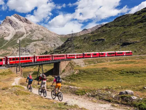 Treno rosso attraversa un ponte su un paesaggio alpino mozzafiato. Tre ciclisti pedalano su un sentiero sottostante, godendosi la vista della ferrovia del Bernina.