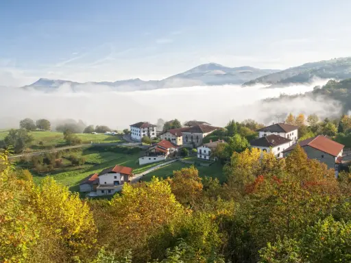 Pueblo en el **valle del Baztan** cubierto por niebla matutina, con casas blancas y tejados rojos entre árboles otoñales.