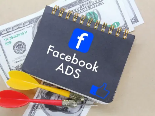 Notatnik z logo Facebook i napisem "Facebook ADS" leży na banknotach dolarowych. Obok znajdują się rzutki, symbolizujące celne reklamy mobilne.