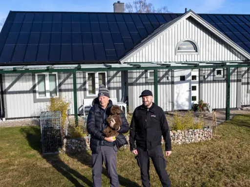 Två män och en hund framför ett hus med solpaneler på taket. Taket har vackra vinklar som ger huset karaktär.