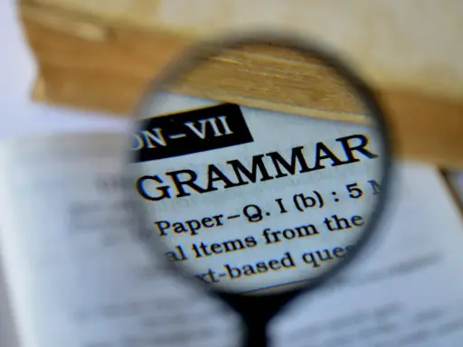 Une loupe examine le mot "GRAMMAR" dans un livre. Pour maîtriser la langue, il faut bien receler la conjugaison.