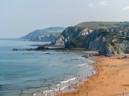Ein belebter Strand mit vielen Menschen, die im Meer schwimmen und surfen, vor einer dramatischen Felsküste. Das Bilbao Meer lädt zum Entspannen ein.