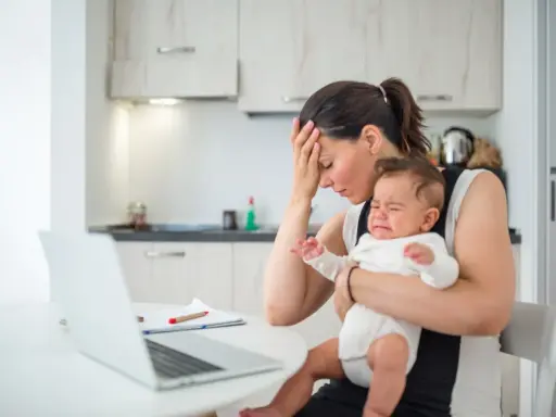 Zmęczona mama z płaczącym niemowlakiem na rękach, zastanawia się, przebodźcowanie niemowlaka ile trwa. Obok laptop i dokumenty.