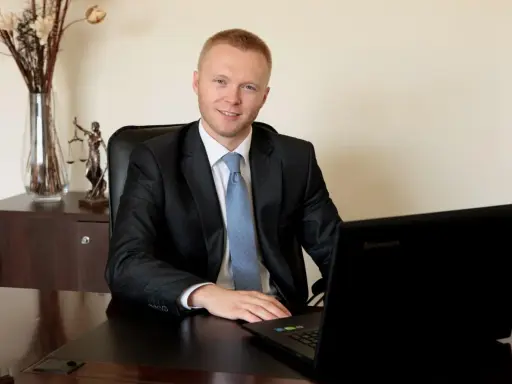 Mężczyzna w garniturze siedzi przy biurku z laptopem, obok statuetka Temidy.