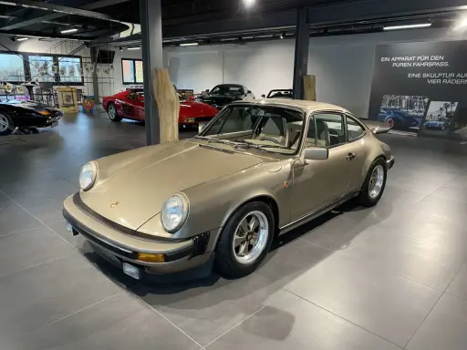 Ein goldener Porsche 911 SC 3.0 steht in einer Ausstellungshalle, flankiert von anderen Sportwagen.