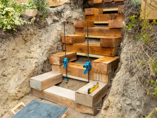 Construction d'un escalier en bastaing dans une pente. Des serre-joints bleus maintiennent les poutres en bois pendant que le niveau à bulle assure la précision.