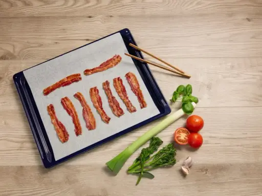 Bacon i ugn på bakplåtspapper, bredvid grönsaker och vitlök. Perfekt för en god måltid.
