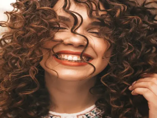 Mujer sonriendo con unos cortes de pelo rizado espectaculares, su cabello castaño cae en cascada alrededor de su rostro.