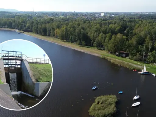 Widok na zbiornik wodny z zaporą i spływającą wodą. Na wodzie kilka łodzi, w tle las i zabudowania. To piękne **chechło** jest idealnym miejscem do wypoczynku.