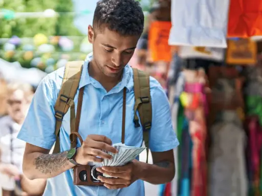 Mężczyzna z plecakiem liczy banknoty dolarów na tle kolorowego straganu.