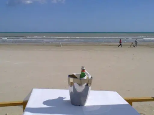 Coppia passeggia sulla spiaggia di Milano Marittima, con un secchiello del ghiaccio e una bottiglia su un tavolo in primo piano.