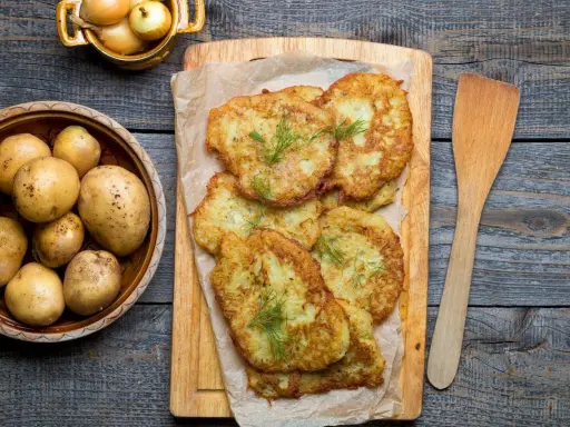 Placki ziemniaczane na desce, obok miski z ziemniakami i cebulą.