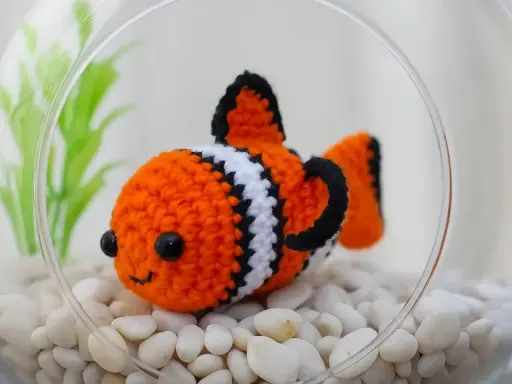 Adorable pez payaso de ganchillo en una pecera con piedras blancas y plantas verdes. ¡Perfecto para patrones de peces de ganchillo!