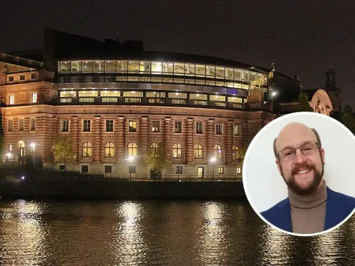 En man ler mot kameran framför Riksdagshuset i Stockholm, en symbol för representativ demokrati.