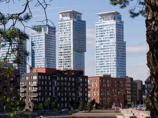 Höga, moderna skyskrapor reser sig bakom tegelbyggnader. En grönskande gren ramar in vyn, som om den undrar när sommaren startar i Sverige.