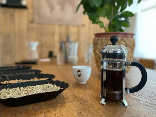 Eine French Press mit frisch gebrühtem Kaffee steht neben einer Tasse und Schalen mit Kaffeebohnen.