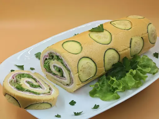 Rotolo di zucchine ripieno di prosciutto e insalata, decorato con fette di zucchina e prezzemolo su un piatto bianco.