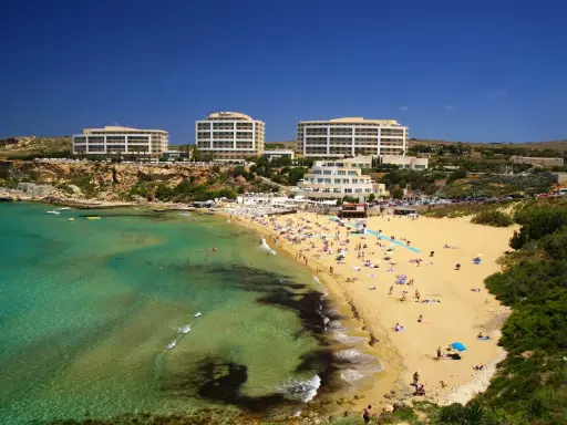 En livlig strand med turkost vatten och gyllene sand vid Golden Bay, Malta. Flera hotell syns i bakgrunden.