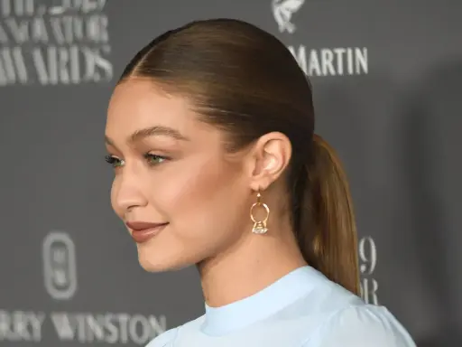 Gigi Hadid mit glatten, **haare nach hinten** gebundenen Haaren und einem dezenten Lächeln. Sie trägt ein hellblaues Oberteil und auffällige Ohrringe.
