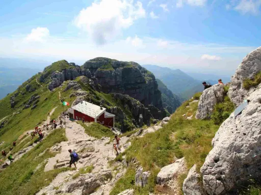 Sentiero panoramico verso il rifugio Lecco, con escursionisti su un crinale erboso e roccioso sotto un cielo azzurro.