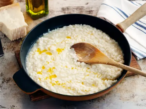 Krämig risotto i panna med en träsked. Bredvid syns parmesanost, olivolja och en rivare. Frågan är om man kan frysa risotto?