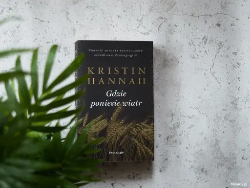 Okładka książki Kristin Hannah "Gdzie poniesie wiatr" na tle szarej ściany, z zielonymi liśćmi rośliny w zbliżeniu.