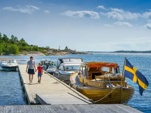 En kvinna och ett barn går på en brygga vid en sjö i Sverige. Båtar ligger förtöjda, en med svensk flagga. En perfekt familjesemester i Sverige.