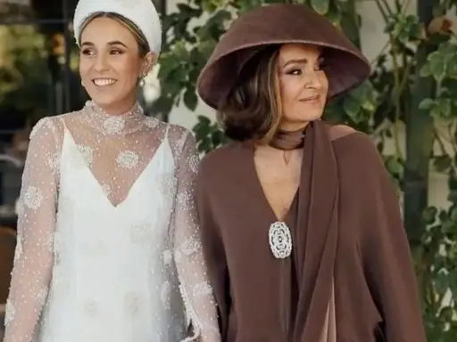 La madre de la novia, elegante con sombrero marrón, acompaña a la radiante novia con su vestido blanco adornado.