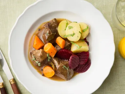 En tallrik med skånsk kalops, mört kött, morötter, potatis och rödbetor. Serveras med en drink och bestick.