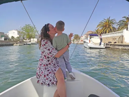 Madre besa a su hijo en un paseo en barco por los canales de Empuriabrava, con palmeras y casas blancas al fondo.