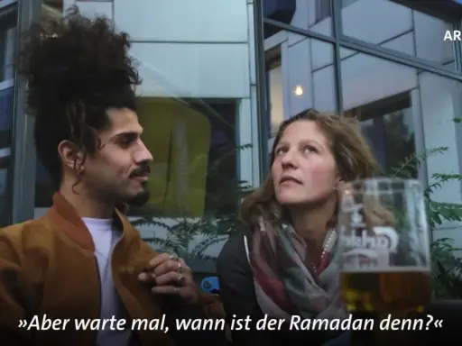 Ein Mann mit vollem Haar und eine Frau sprechen. Er fragt: "Aber warte mal, wann ist der Ramadan denn?" Die Fastenzeit im Islam ist ein wichtiges Thema.