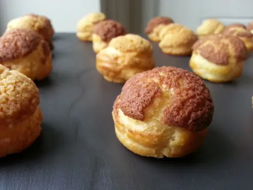 Une rangée de choux craquelins dorés et croustillants, certains avec un dessus brun, prêts à être dégustés.