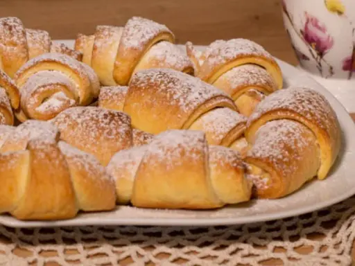Talerz pełen puszystych, posypanych cukrem pudrem rogalików krucho drożdżowych. Idealne na śniadanie lub deser.