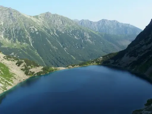 Górski krajobraz z jeziorem i stromymi zboczami. W oddali widać pasmo górskie, które przypomina najwyższy szczyt w Polsce.