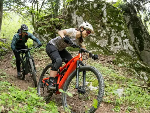 Dwie osoby na rowerach zjazdowych, jeden pomarańczowy rower bez amortyzatora z przodu, jadą po leśnym szlaku.