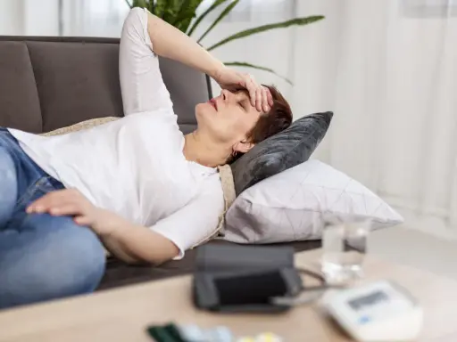 Donna sdraiata sul divano con la mano sulla fronte, visibilmente affaticata e con la paura di svenire. Sul tavolino, un misuratore di pressione.