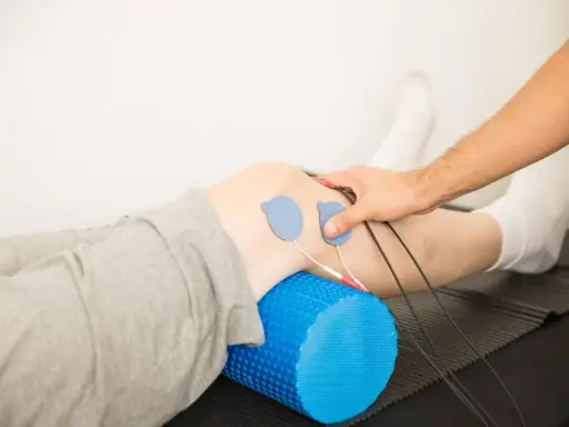 Rehabilitacja kolana w domu: elektrody na skórze, wałek do masażu pod nogą, ręka terapeuty.