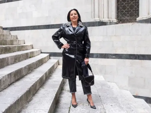 Donna con un audace outfit rock: trench in ecopelle nera, jeans scuri, décolleté borchiate e borsa a tracolla.