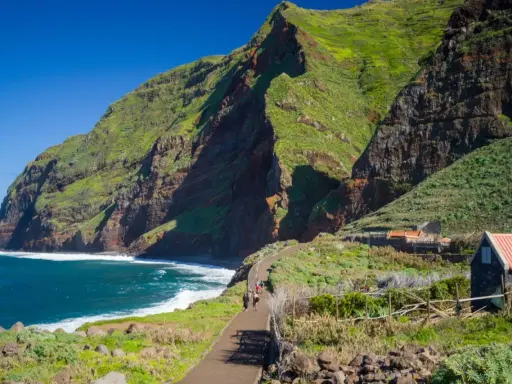 Grüne Berge und rote Felsen prägen die Küste der Madeira Inselgruppe. Ein Weg führt zu kleinen Häusern.