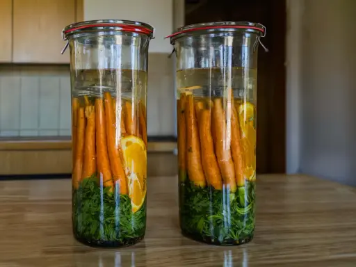 Zwei Gläser mit Karotten, Kräutern und Orangenscheiben in Salzlake zum Fermentieren.
